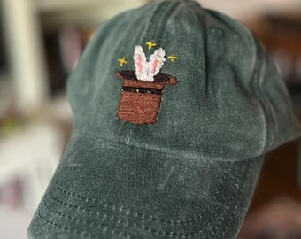 Könnte beinhalten: Dunkelgrüne Baseballkappe mit einem gestickten Design eines braunen Zauberhuts und eines weißen Kaninchens, das daraus hervorkommt. Das Design wird durch gelbe Sternchen akzentuiert. Die Kappe hat einen gebogenen Schirm und einen Stoffriemen.