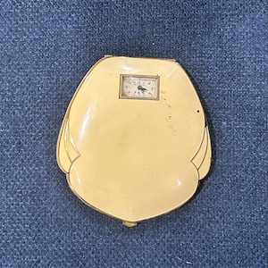 May include: A vintage compact with a small clock. The compact is a light cream color with gold trim and a small rectangular clock face at the top. The clock has black numbers and hands.