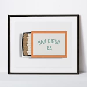 Peut inclure: Impression encadrée représentant une boîte d'allumettes ouverte avec des allumettes et le texte "SAN DIEGO CA" en bleu clair. Le cadre est noir, sur fond blanc.