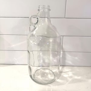 May include: A clear glass jug with a handle. The jug has a wide base and a narrow neck, and is designed for holding liquids. The jug is empty and sits on a white surface, with a white tile background.