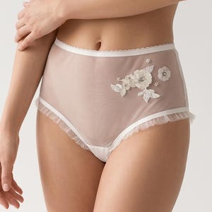 Op de afbeelding: High-waisted slip in een doorschijnende, nude stof met witte rand en een gerimpelde rand. De voorkant heeft een geborduurd bloemmotief in wit. Een delicaat en vrouwelijk lingeriestuk.