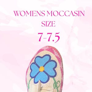 May include: A cream-coloured moccasin with a beaded blue flower design. The moccasin has a pink and white beaded border. The text "WOMENS MOCCASIN SIZE 7-7.5" is displayed in pink.