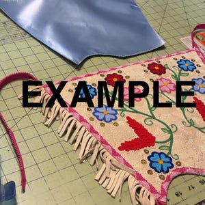 K&ouml;nnte beinhalten: Ein hellblaues Satin-Stoffst&uuml;ck und ein Perlenhandwerk mit floraler Stickerei. Das Perlenst&uuml;ck hat einen rosa Rand und Fransen. Das Wort "EXAMPLE" ist schwarz am unteren Bildrand aufgedruckt.