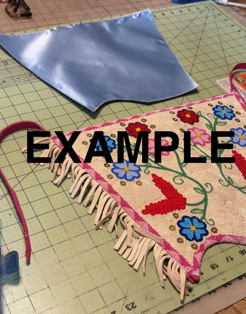 K&ouml;nnte beinhalten: Ein hellblaues Satin-Stoffst&uuml;ck und ein Perlenhandwerk mit floraler Stickerei. Das Perlenst&uuml;ck hat einen rosa Rand und Fransen. Das Wort "EXAMPLE" ist schwarz am unteren Bildrand aufgedruckt.