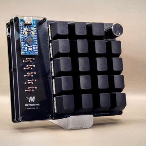 May include: A black mechanical keyboard with a small circuit board and a rotary encoder. The keyboard has a 5x5 key layout with square keycaps. The text "MATRIXE PRO" is visible on the side.