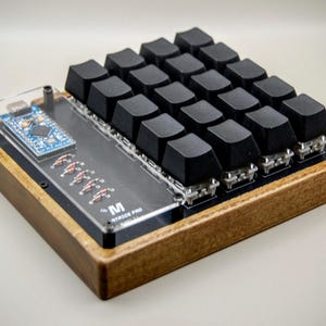 Peut inclure: Un clavier mécanique personnalisé avec des touches noires et un cadre en bois. Le clavier a un dessus en acrylique transparent et une petite carte de circuit imprimé avec des composants électroniques. Le texte "M" et "MATRIX PRO" est visible.