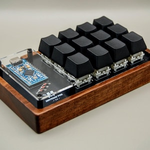 May include: A custom macro pad with a wooden base and black keycaps. The pad has a clear acrylic top with a circuit board and the text "MATRIX PRO". The wooden base is finished in a dark brown colour.