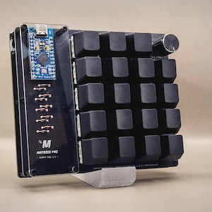 Peut inclure: Un clavier Matrix Pro noir avec une carte de circuit imprimé bleue et un bouton argenté. Le clavier a une grille de touches carrées noires et est monté sur un support en plastique transparent. Le texte "M MATRIX PRO" est visible.