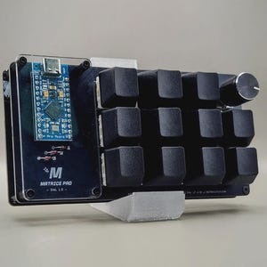 May include: A black Matrice Pro macro pad with 12 keycaps, a rotary encoder, and a blue circuit board. The pad has a clear acrylic cover and a white support base. The text "Matrice Pro - DNL 1.0" is printed on the black surface.