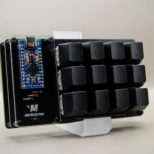 May include: A black Matrice Pro 2.0 keyboard with a blue circuit board and black keycaps. The keyboard has a rectangular design and is supported by a white stand. The text "Matrice Pro - 2.0" is visible.