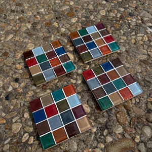 Puede incluir: Cuatro posavasos cuadrados con un diseño de mosaico. Cada posavasos presenta una cuadrícula de pequeñas baldosas de colores en tonos de rojo, azul, marrón y verde, sobre una lechada blanca. Los posavasos están dispuestos sobre una superficie de piedra texturizada.