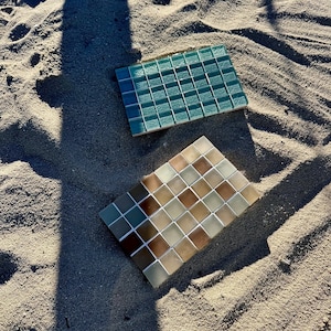May include: Two rectangular mosaic tile sheets. One sheet features a grid of square glass tiles in varying shades of teal and blue. The second sheet consists of a grid of square tiles in a gradient of warm beige, tan, and brown earth tones.