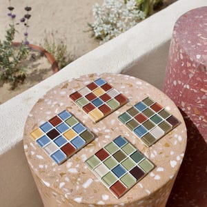 Könnte beinhalten: Ein Set aus drei quadratischen Untersetzern, die jeweils aus einem Gitter von sechzehn kleinen Glasfliesen in verschiedenen Farben wie Blau, Rot, Gelb, Grün und Beige bestehen. Die Untersetzer haben eine glatte, reflektierende Oberfläche.