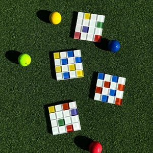 May include: An overhead shot shows a green surface with five square mosaic tile coasters in a grid pattern, each with a different color scheme. Scattered around are four golf balls in yellow, green, blue, and red.