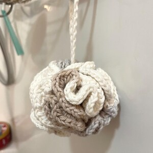 May include: A round, crocheted bath sponge featuring a layered, ruffled design in a neutral color palette of cream, beige, and light gray. The item includes a matching crocheted loop at the top for hanging, constructed from soft yarn.