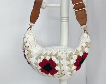 May include: A white crochet bag with a brown adjustable strap. The bag features a floral pattern with red and black accents. The bag is hanging on a white stand.
