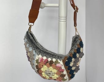 May include: A crocheted crescent-shaped bag with a brown adjustable strap. The bag features a patchwork design in various colors, including gray, pink, yellow, and blue. The strap is brown with gold-colored hardware.