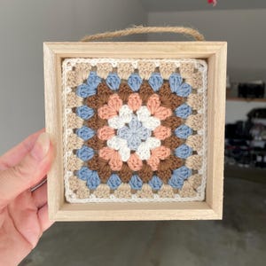 May include: A square crochet granny square wall hanging framed in light-colored wood. The textile features a geometric pattern in shades of light blue, white, peach, and brown, with a jute twine loop attached to the top for hanging purposes.