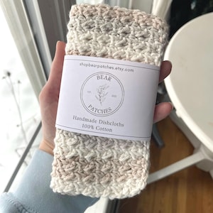 May include: A rectangular, crocheted dishcloth made of 100% cotton in variegated shades of cream, beige, and white. A white paper band wraps around the center, featuring the text "shopbearpatches.etsy.com", "BEAR PATCHES", and "Handmade Dishcloths".