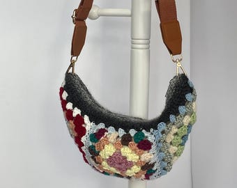 May include: A handmade crochet shoulder bag in a crescent shape. The bag features a colorful granny square design with shades of red, green, yellow, and blue. It has a brown adjustable strap with gold-tone hardware.