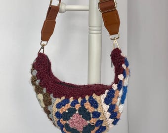 May include: A crescent-shaped, handmade crochet bag with a burgundy interior and a colorful, geometric pattern on the exterior. The bag has a brown adjustable strap with gold-colored hardware.