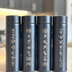 May include: Four black insulated water bottles with silver Unown symbols. Each bottle has a screw-on lid and a cylindrical shape. The Unown symbols are arranged vertically on the bottles. The bottles are displayed on a light-colored surface.