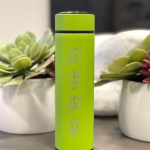 May include: A neon green insulated water bottle with a black cap and a series of cartoon character outlines. The bottle is cylindrical and appears to be made of metal. The background includes white planters with green and red plants.