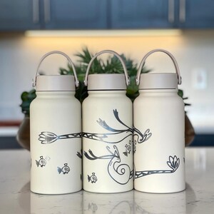 Könnte beinhalten: Drei cremefarbene Wasserflaschen mit silbernen Illustrationen von Fischen und skurrilen Designs. Jede Flasche hat einen hellbraunen Deckel und Griff. Die Flaschen stehen auf einer weißen Arbeitsplatte mit verschwommenem Hintergrund.