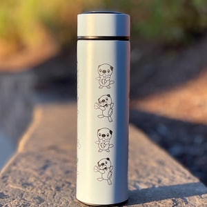 May include: A light blue insulated water bottle featuring a repeating pattern of a cartoon otter holding a seashell. The bottle has a black rim and a matching light blue lid. The background is blurred.