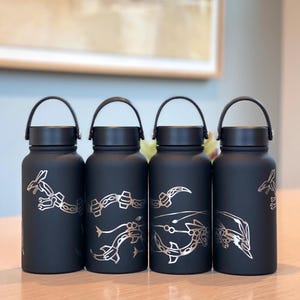 Puede incluir: Cuatro botellas de agua negras con diseños de dragones plateados estilizados. Cada botella tiene un asa y una tapa negras. Las botellas son cilíndricas y están sobre una superficie de madera clara.