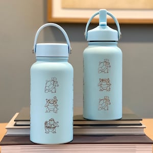 May include: Two pale blue water bottles with matching lids and handles. Each bottle has brown cartoon turtle illustrations. The cylindrical bottles are on a stack of books. The background includes a framed picture and a light-coloured wall.