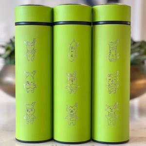 May include: Three lime green insulated water bottles with black accents. Each bottle features a series of silver cartoon character illustrations. The bottles are cylindrical and appear to be made of metal. The background is blurred.