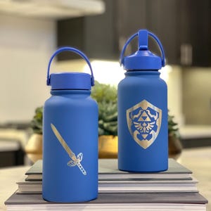 May include: Two blue water bottles with silver graphics. One bottle features a sword design, and the other displays a shield emblem. Both bottles have blue handles and lids. They are cylindrical and sit on a stack of books.