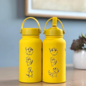 May include: Two yellow water bottles with black cartoon animal illustrations. Each bottle has a yellow handle and lid. The bottles are cylindrical and appear to be made of metal. The background is a light blue wall and a framed picture.