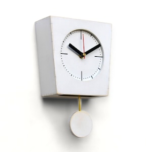 Puede incluir: Un reloj de pared blanco de forma cuadrada con una esfera circular. El reloj tiene manecillas negras, un segundero rojo y un péndulo dorado. El reloj está montado en una pared blanca.