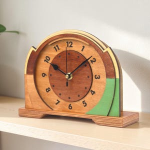 May include: Wooden Art Deco style clock with a light brown, medium brown, and green color scheme. The clock has a circular face with black hands and numbers. The clock is approximately 7 inches wide and 6 inches tall.