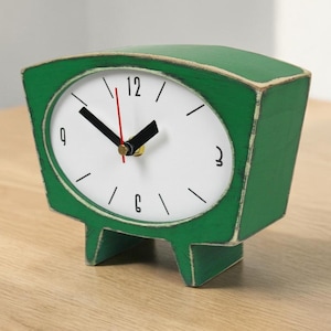 May include: A green table clock with a distressed finish and a white face. The clock features black hour and minute hands, a thin red second hand, and black numerals at the 12, 3, 6, and 9 positions. It stands on two short, wide legs.