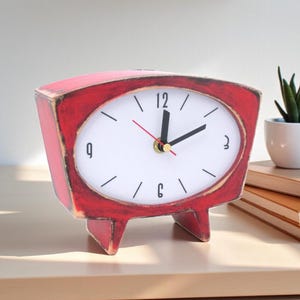 May include: A red, trapezoidal-shaped analog table clock with a distressed finish. The clock face is white with black numerals at the 3, 6, 9, and 12 positions, black hour and minute hands, and a thin red second hand. It stands on two short legs.