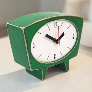 May include: A green table clock with a distressed finish and a white oval face. The clock features black hour and minute hands, a red second hand, and black numerals at the 12, 3, 6, and 9 positions. The clock is supported by two small feet.