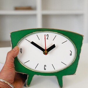 May include: A green wooden table clock with a white oval face, black hour and minute hands, and a thin red second hand. The clock features black numerals at the 12, 3, 6, and 9 positions, with simple black lines for the remaining hour markers.