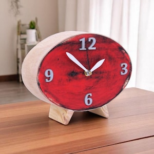 Könnte beinhalten: Eine ovale Holzuhr mit rotem Zifferblatt, weißen Zeigern und Ziffern. Die Uhr wird von einem kleinen Holzständer getragen. Das Zifferblatt zeigt die Zahlen 12, 3, 6 und 9.