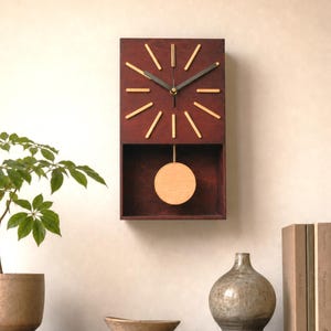 May include: A rectangular wall clock with a dark brown wooden frame, featuring a circular light wood pendulum swinging in an open lower compartment. The clock face has twelve light wood hour markers and two dark metal hands for timekeeping.