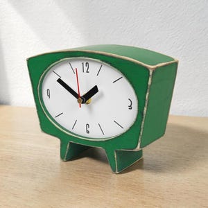 Può includere: Un orologio da scrivania verde in stile retrò, con quadrante bianco e numeri neri. L'orologio ha lancette nere e rosse ed è appoggiato su una superficie di legno. Il corpo dell'orologio ha una forma squadrata con la parte superiore curva e piedini.