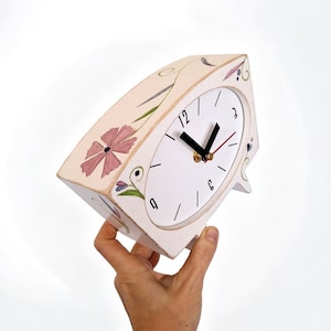 Puede incluir: Reloj blanco con detalles florales y agujas negras. La esfera del reloj tiene números romanos negros y está configurada a las 3:00. El reloj está sobre una mesa.