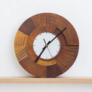 Op de afbeelding: Een ronde houten klok met een witte wijzerplaat en zwarte wijzers. De lijst van de klok is gemaakt van verschillende bruintinten, met een gesneden geometrisch ontwerp. De klok wordt tentoongesteld op een lichtgekleurde houten plank.