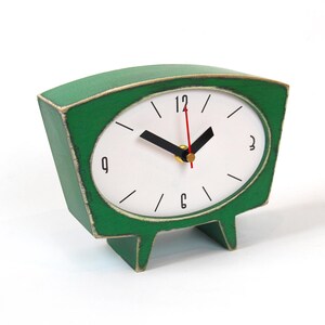 May include: A green table clock with a trapezoidal wooden frame featuring a distressed finish. The white oval clock face displays black numerals at 12, 3, 6, and 9, with black hour and minute hands and a thin red second hand on a central gold pin.