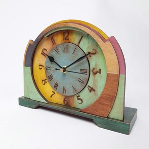 May include: A wooden mantel clock with a multi-coloured face and a green base. The clock face has Roman numerals and black hands. The clock is painted in shades of blue, yellow, green, brown, and pink. The clock measures approximately 25 cm in height.