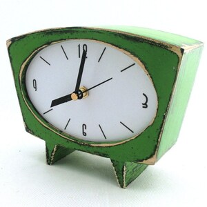 May include: A trapezoidal table clock with a distressed green finish and a white face. The clock features black hour markers, black hands, and numerals at the 12, 3, 6, and 9 o'clock positions. It stands on two small, integrated green feet.