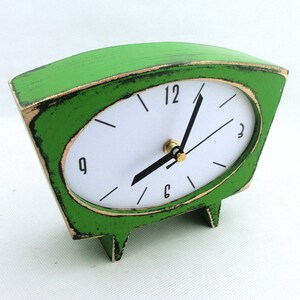 May include: A trapezoidal table clock with a distressed bright green finish and black edges. The clock face is white with black numerals at 12, 3, 6, and 9, featuring black hour and minute hands and a gold-toned center pin mechanism.