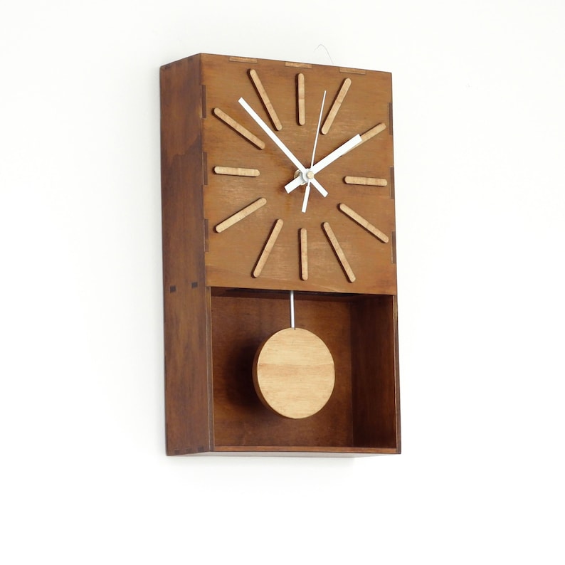 Puede incluir: Un reloj de pared rectangular hecho de madera marr&oacute;n oscura con un p&eacute;ndulo de madera marr&oacute;n m&aacute;s claro. La esfera presenta doce marcadores de madera delgados y claros para las horas y dos manecillas blancas. El p&eacute;ndulo es un disco circular.